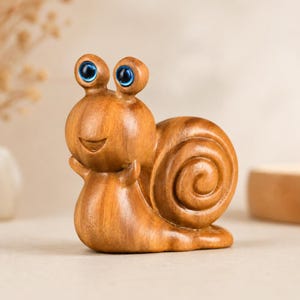 Puede incluir: Figura de caracol de madera con una cara sonriente y ojos azules. La concha del caracol tiene un diseño en espiral. La figura está hecha de madera marrón claro y tiene un acabado liso. El caracol está de pie.