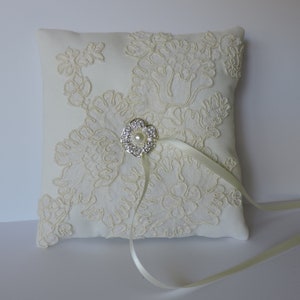 May include: A white satin ring pillow with a delicate lace design and a silver flower brooch with a pearl center. The pillow has a satin ribbon attached.