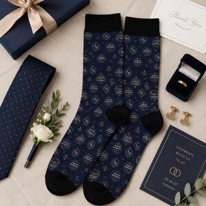 May include: Navy blue socks with black heels and toes, featuring a gold pattern of initials, rings, and text. A navy blue tie with white polka dots, a gift box, and a card are also visible.