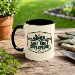 Op de afbeelding: Crèmekleurige keramische mok met een zwarte binnenkant en oor. De mok heeft een groen en goud ontwerp met een grasmaaierafbeelding en de tekst "YARD SHIFT SUPERVISOR". De mok staat op een houten oppervlak.