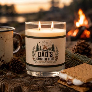 May include: A clear glass double-wick candle with a cream-colored wax surface. The label features a graphic of a campfire, pine trees, and the text DAD'S CAMPFIRE RESET, PINE, SMOKE, MEMORIES, SOY WAX CANDLE. The candle is lit with two visible flames.