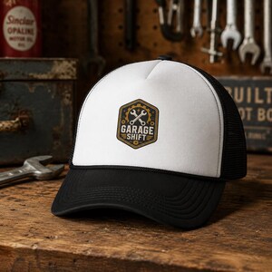 Pode incluir: Boné de caminhoneiro preto e branco com aba curva. O painel frontal é branco, exibindo um emblema dourado e preto com as palavras "GARAGE SHIFT" e chaves cruzadas. O boné tem uma parte traseira de malha preta e fecho de pressão.