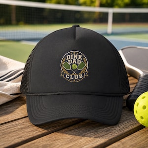 May include: Black trucker hat with a mesh back and a curved peak. The front displays a circular emblem with the text "Dink Dad Club" in gold and white, featuring pickleball paddles and a ball. The hat is on a wooden surface.