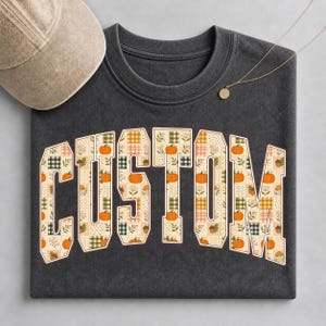 Könnte beinhalten: Ein anthrazitfarbenes T-Shirt mit Rundhalsausschnitt und dem Wort CUSTOM in großen Blockbuchstaben. Die Buchstaben sind mit einem Patchwork-Muster aus orangefarbenen Kürbissen, grünem Karomuster und Herbstlaub gefüllt. Eine beige Kappe und eine Goldkette.
