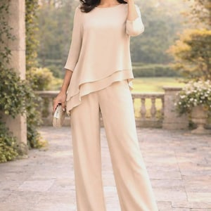 May include: A beige two-piece outfit consisting of a layered top with three-quarter sleeves and wide-leg pants. The outfit is paired with a small, sparkly clutch. The ensemble is suitable for formal events.