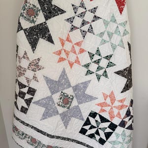 May include: A white quilt featuring a variety of star-shaped blocks in assorted floral and botanical prints. The colors include black, red, coral, sage green, and light blue. The fabric is stitched with a swirling, continuous line quilting pattern.