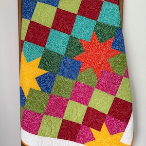May include: A patchwork quilt featuring a grid of multicolored squares in shades of red, blue, green, yellow, and pink, all with a fine white speckled pattern. The design incorporates large, solid-colored eight-pointed stars in orange and yellow.