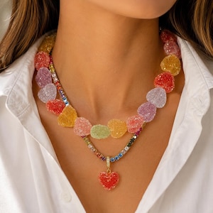 Könnte beinhalten: Eine mehrfarbige herzförmige Halskette mit einer Regenbogenfarbenen Edelsteinkette und einem roten Herzanhänger. Die Halskette hat eine Vielzahl von Farben, darunter Rot, Gelb, Grün und Lila. Die Halskette wird über einem weißen Hemd getragen.