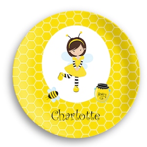 May include: A yellow ceramic plate with a honeycomb pattern. The center of the plate features a cartoon girl dressed as a bee, holding a jar of honey that says "Honey". The plate also has the name "Charlotte" written in black letters.