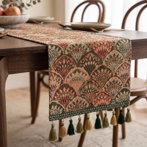 May include: A rectangular table runner featuring a repeating fan-shaped pattern in shades of terracotta, sage green, and cream. The fabric has a woven texture and is finished with a decorative border and a row of hanging tassels in green and gold.