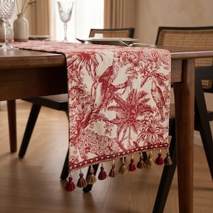 May include: A rectangular table runner featuring a red and cream toile pattern with tropical birds, palm trees, and foliage. The fabric edge is finished with a decorative trim and a series of hanging tassels in matching red and muted gold tones.