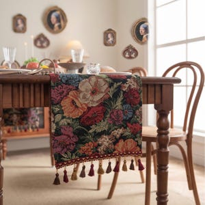 Peut inclure: Un chemin de table rectangulaire orné d'un motif floral tissé avec des fleurs rouges, orange, roses et bleues sur fond noir. Le chemin de table est agrémenté d'une bordure décorative rouge foncé et d'une rangée de pompons marron et dorés.