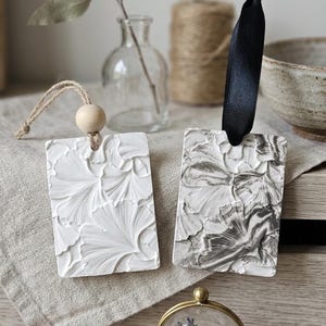 Peut inclure: Deux ornements rectangulaires à suspendre avec des motifs de feuilles de ginkgo en relief. L'un est blanc uni avec une cordelette en jute et une perle en bois, l'autre présente une finition marbrée noire et blanche avec un ruban en satin noir.