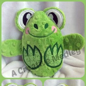 May include: A green frog plush toy with embroidered details. The frog is wearing a blue and green blanket. The frog is smiling and has big eyes.