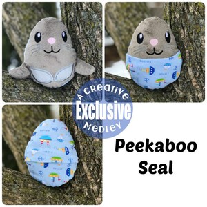 May include: A gray and white plush seal toy with a blue and white fabric covering the bottom half of the body. The fabric has a nautical theme with sailboats, anchors, and the words "Little Sailor".