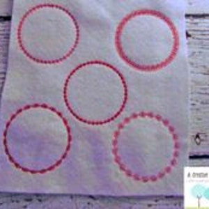 May include: White fabric with five embroidered red circles of varying designs. The circles are outlined with different stitch patterns, including dotted and scalloped edges. The fabric has a slightly textured appearance.