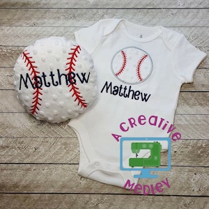 Puede incluir: Un body blanco para bebé con un gráfico de béisbol rojo y blanco y el nombre "Matthew" bordado en azul. También se muestra un balón de béisbol de felpa blanco con el nombre "Matthew" bordado en azul.