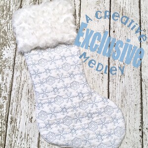 May include: White quilted Christmas stocking with a faux fur cuff and a blue snowflake pattern. The stocking is on a whitewashed wood background. The text "A Creative Exclusive Medley" is in blue letters on the background.