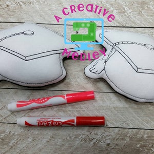 May include: Two white felt graduation caps with black outlines and a tassel. The caps are lying on a wooden surface with two red markers.