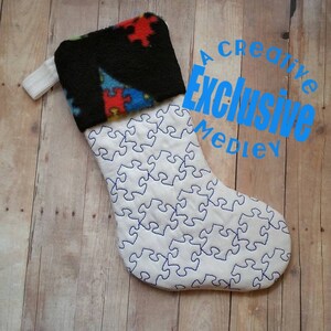 May include: White Christmas stocking with a blue puzzle piece design. The stocking has a black fleece cuff with a red, blue, and white puzzle piece design.