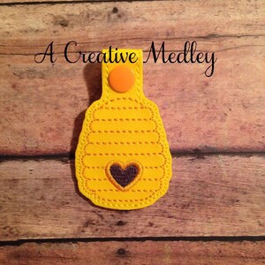 May include: A yellow felt keychain shaped like a honeycomb with a small black heart in the center. The keychain has a button closure and the text "A Creative Medley" is visible in the background.