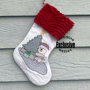 May include: White Christmas stocking with a red faux fur cuff. The stocking features an embroidered design of a white bear holding a green Christmas tree with presents underneath. The text "A Creative Exclusive Medley" is embroidered in a circle on the stocking.