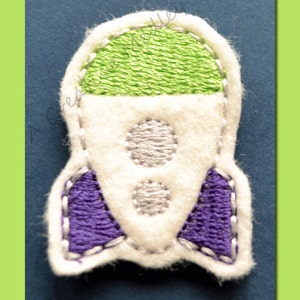 Puede incluir: Un cohete de fieltro blanco con detalles de fieltro morado y verde. El cohete tiene dos círculos plateados en el cuerpo.