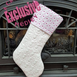 May include: White Christmas stocking with a pink quilted design and a pink and white patterned cuff. The stocking is hanging on a black fireplace.