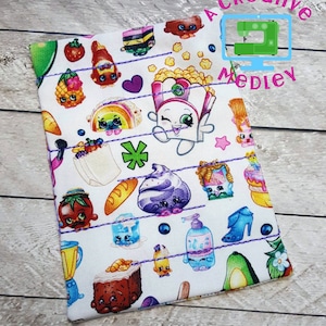 Könnte beinhalten: Eine weiße Stofftasche mit einem bunten Muster aus Comicfiguren und Lebensmitteln. Die Tasche hat einen lilafarbenen Stich entlang der Kanten. Der Text "Creative Medley" ist in der oberen rechten Ecke der Tasche aufgedruckt.