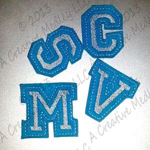 May include: Four felt letters, S, M, C, and V, in a blue color with a white outline. The letters are stitched together with a white thread.
