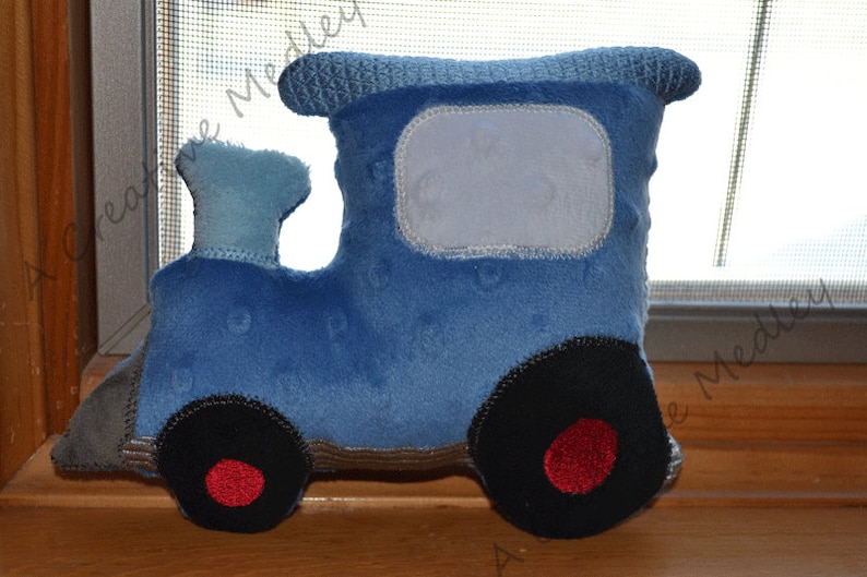 May include: A blue plush toy train with a grey and brown base. The train has a large window and two black wheels with red centres.