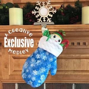 May include: A blue and white quilted stocking with snowflake designs hangs from a silver snowflake ornament. The stocking is filled with candy canes and a small doll with a red hat.