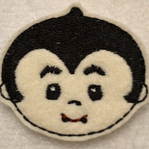 May include: A white felt embroidered patch of a cartoon vampire with black hair and a red mouth.