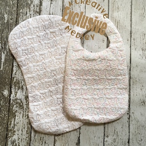 May include: Two white quilted baby bibs with a leaf pattern. The bibs are made of soft fabric and have a comfortable fit. The bibs are embroidered with a leaf pattern in brown, pink, and blue thread. The bibs are perfect for keeping baby's clothes clean during mealtime.