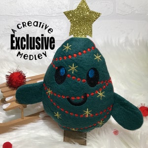 May include: A green felt Christmas tree plush toy with a smiling face, red and gold embroidery, and a gold glitter star on top. The plush toy is sitting on a wooden sled.