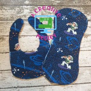 May include: Two blue and white fabric bibs with a space theme. One bib features a cartoon astronaut in a spacesuit holding a pizza. The other bib features a cartoon astronaut in a spacesuit holding a pizza and the text "To Infinity and Beyond".