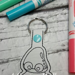 May include: A white leather keychain with a black outline of a cartoon train. The train has two wheels and a small window. The keychain has a light blue button on the top and a silver key ring.