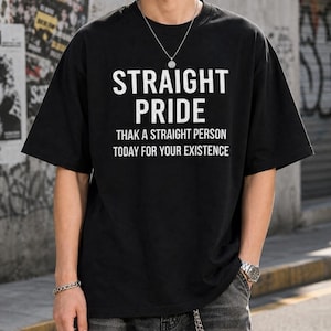 May include: A black short-sleeved t-shirt featuring white text that reads STRAIGHT PRIDE THAK A STRAIGHT PERSON TODAY FOR YOUR EXISTENCE. The shirt has a crew neckline and a loose fit, worn with a silver chain necklace and dark denim jeans.