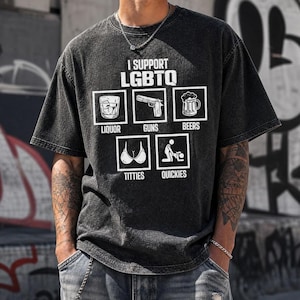 Peut inclure: Un t-shirt gris anthracite à manches courtes avec le texte I SUPPORT LGBTQ au-dessus de cinq icônes carrées représentant un verre d'alcool, une arme de poing, une chope de bière, un soutien-gorge et une silhouette sexuelle, étiquetés LIQUOR, GUNS, BEERS, TITTIES, QUICKIES.
