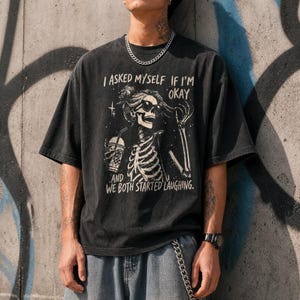 May include: Dark gray t-shirt with a graphic of a skeleton wearing sunglasses and holding a drink. The text on the shirt reads, "I asked myself if I'm okay, and we both started laughing."