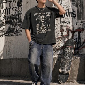 May include: A black graphic t-shirt with the text "I ASKED MYSELF IF I'M OKAY AND WE BOTH STARTED HING." The shirt is paired with blue jeans and a skateboard. The background features graffiti and posters.