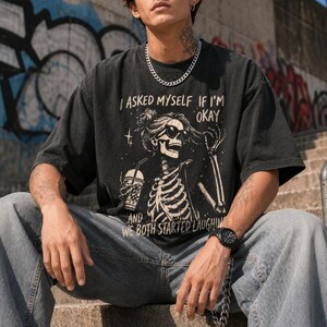 May include: Dark gray t-shirt with a skeleton graphic wearing sunglasses and holding a drink. The text on the shirt reads "I ASKED MYSELF IF I'M OKAY AND WE BOTH STARTED LAUGHIN". The shirt is paired with light blue jeans.