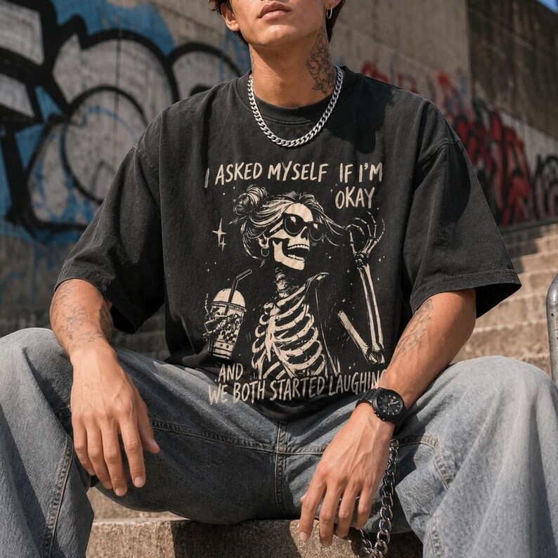 May include: Dark gray t-shirt with a skeleton graphic wearing sunglasses and holding a drink. The text on the shirt reads "I ASKED MYSELF IF I'M OKAY AND WE BOTH STARTED LAUGHIN". The shirt is paired with light blue jeans.