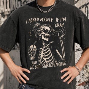 May include: Dark gray t-shirt with a skeleton graphic wearing sunglasses and holding a drink. The text on the shirt reads, "I asked myself if I'm okay, and we both started laughing."