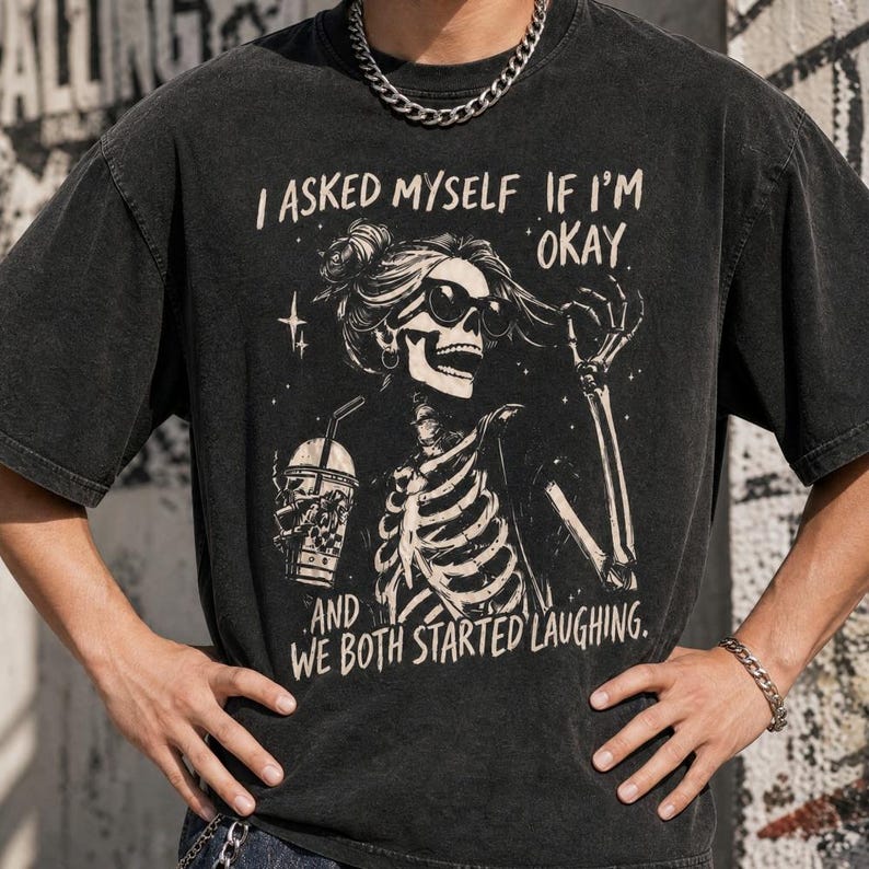 May include: Dark gray t-shirt with a skeleton graphic wearing sunglasses and holding a drink. The text on the shirt reads, "I asked myself if I'm okay, and we both started laughing."