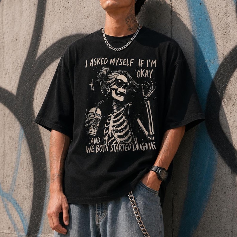 May include: Black t-shirt with a skeleton graphic wearing sunglasses and holding a beverage. The text on the shirt reads "I ASKED MYSELF IF I'M OKAY AND WE BOTH STARTED LAUGHING."