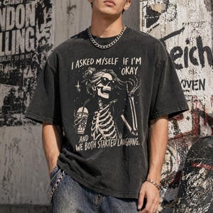 May include: Dark gray t-shirt with a graphic of a skeleton wearing sunglasses and holding a drink. The text on the shirt reads, "I ASKED MYSELF IF I'M OKAY AND WE BOTH STARTED LAUGHING."