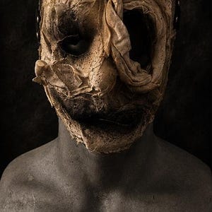 May include: A full-head mask featuring a textured, beige, fabric-wrapped surface with dark, distressed detailing. The mask has deep, hollow eye sockets and a wide, dark mouth opening, displayed on a grey, textured, human-torso-shaped mannequin bust.