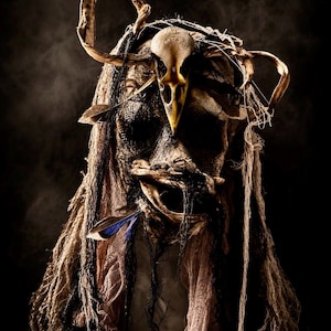 May include: A decorative mask featuring a central bird skull, draped in layers of distressed, frayed tan and black fabric. The mask is adorned with natural feathers, including a prominent blue-tipped feather, and textured, weathered netting.