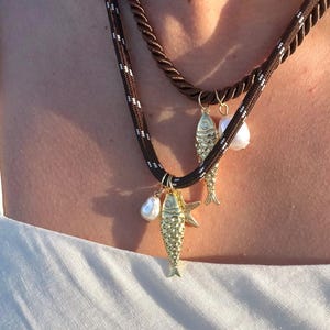 May include: Two layered necklaces with brown and black rope-like cords. The lower necklace features two gold fish pendants and a pearl. The upper necklace is a solid brown rope. The necklaces are worn over a white top.